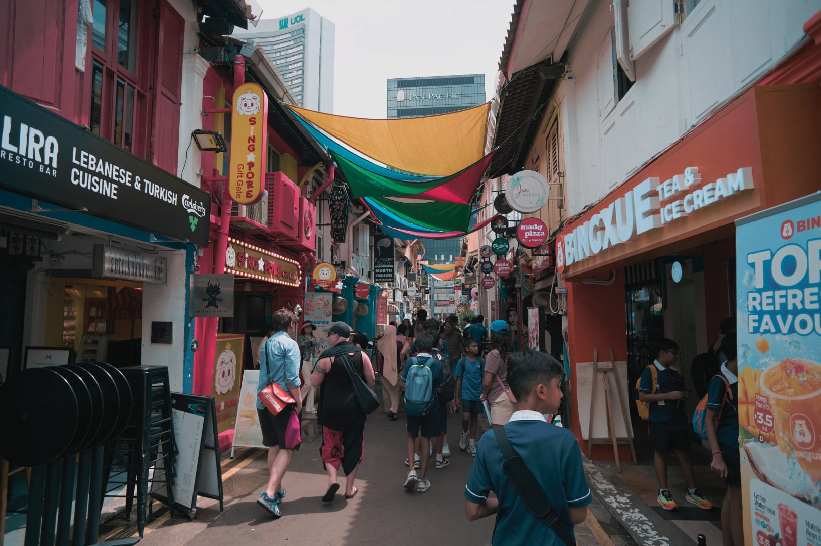 A narrow, colorful alleyway in Singapore, bustling with people walking and lined with vibrant businesses like LIRA Lebanese Cuisine and BINGXUE Tea & Ice Cream.