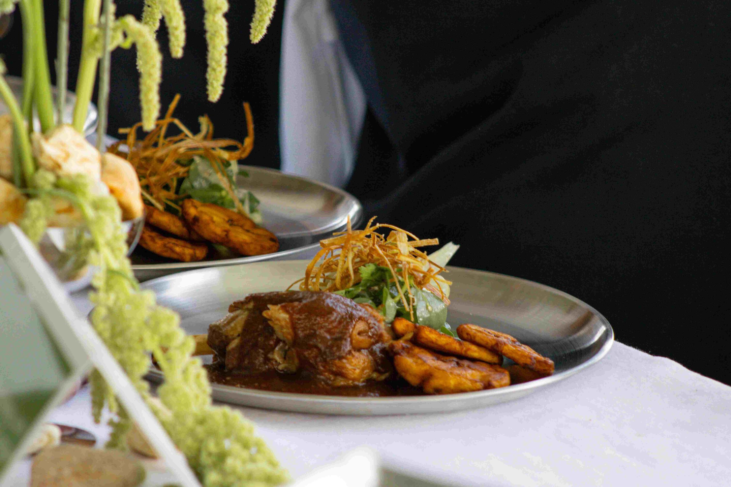 Two plates of beautifully plated food, featuring a meat dish in a rich brown sauce, several slices of fried plantain, and a small green salad topped with thin, crispy fried strands, all served on silver plates on a white tablecloth. A waiter in a black uniform is visible in the background.