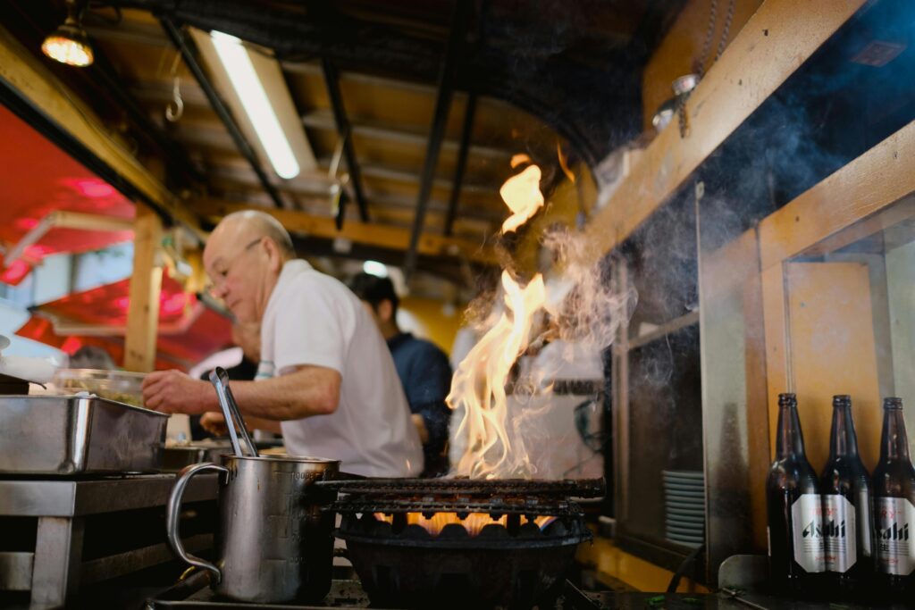 Bright orange flames and swirling smoke rise dramatically from a grill, taking center stage in this bustling kitchen scene. In the background, a chef diligently prepares food near a row of Asahi beer bottles, creating an atmosphere typical of a lively izakaya or street food stall.