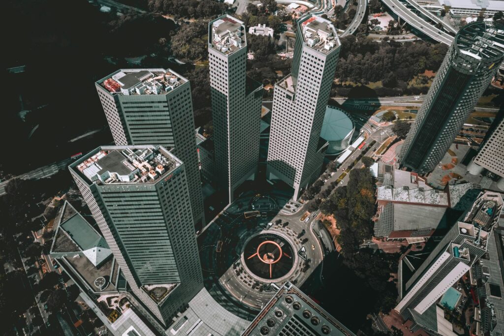 This high-angle aerial photograph captures a cluster of modern skyscrapers arranged around a large, central traffic roundabout. At the heart of the circle lies a distinctive ring-shaped fountain structure, anchoring the busy urban landscape of roads and greenery.