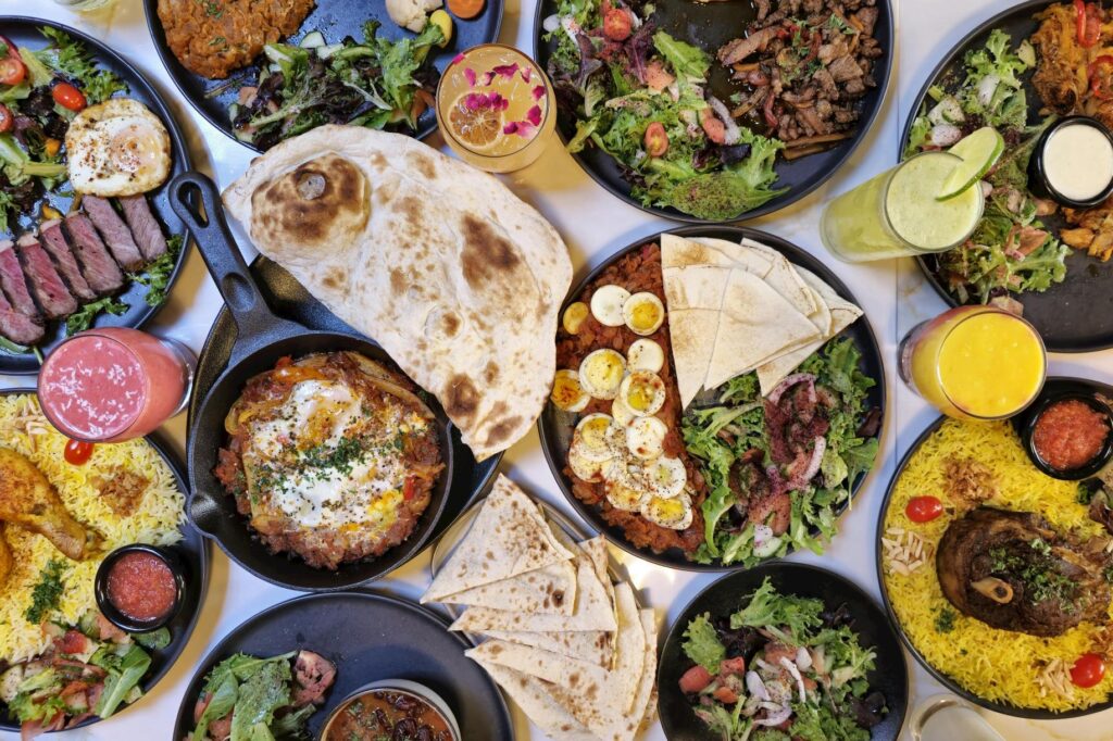 This vibrant top-down photograph showcases a lavish Middle Eastern feast featuring dishes like shakshuka, grilled meats, yellow rice, and fresh green salads served on black plates. Accompanied by large pieces of flatbread and colorful smoothies, the spread offers a rich variety of textures and flavors ideal for communal dining.