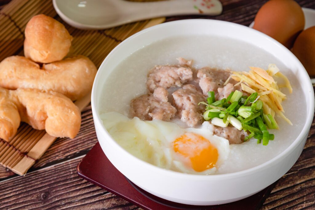 This image features a comforting white bowl of rice porridge, or congee, generously topped with seasoned pork chunks, a soft-poached egg, julienned ginger, and fresh green onions. To the left, a serving of golden-brown fried dough sticks rests on a bamboo mat, completing this classic and savory Asian breakfast arrangement.