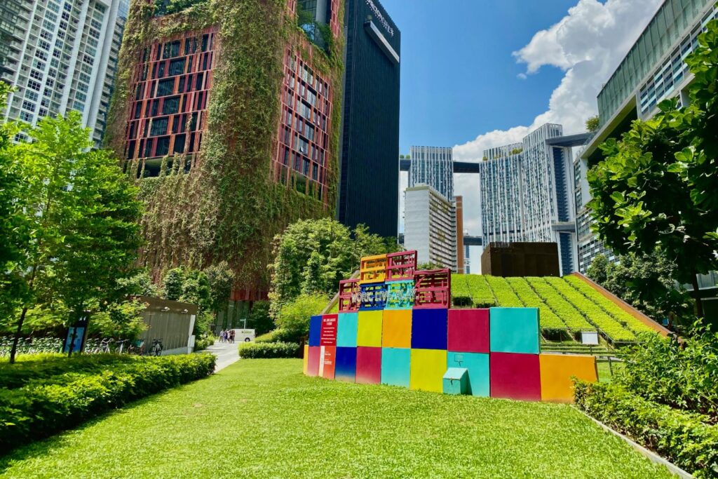 This vibrant urban landscape features a striking high-rise building covered in vertical gardens rising above a lush, sunny park. In the foreground, a playful installation of bright, multicolored cubes sits on a manicured lawn, emphasizing the harmonious blend of modern architecture and green space.