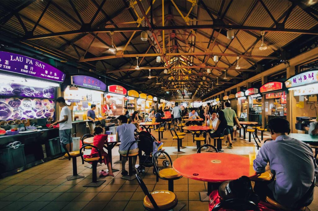 Hawker Centre interior featuring rows of food stalls, high arched ceilings, round tables, and diners enjoying local street food.