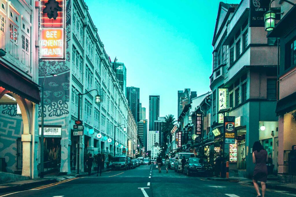 Nighttime city street on Tanjong Tagar featuring neon storefronts, cultural signage, pedestrians, and a contrast of low-rise shops and tall towers.