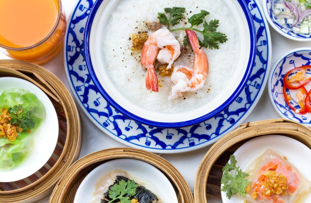This image features a top-down view of an oriental breakfast set, centered around a bowl of shrimp congee garnished with cilantro and black pepper. The meal is complemented by several bamboo steamer baskets containing various dim sum, along with small side dishes of chilies, aromatics, and a glass of orange juice.