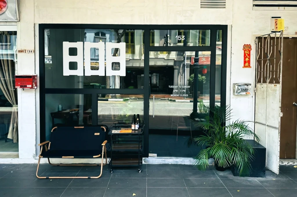 Front entrance of 808 Eating House with large glass windows, minimalist signage, a small bench, potted plants, and a side table near the doorway.
