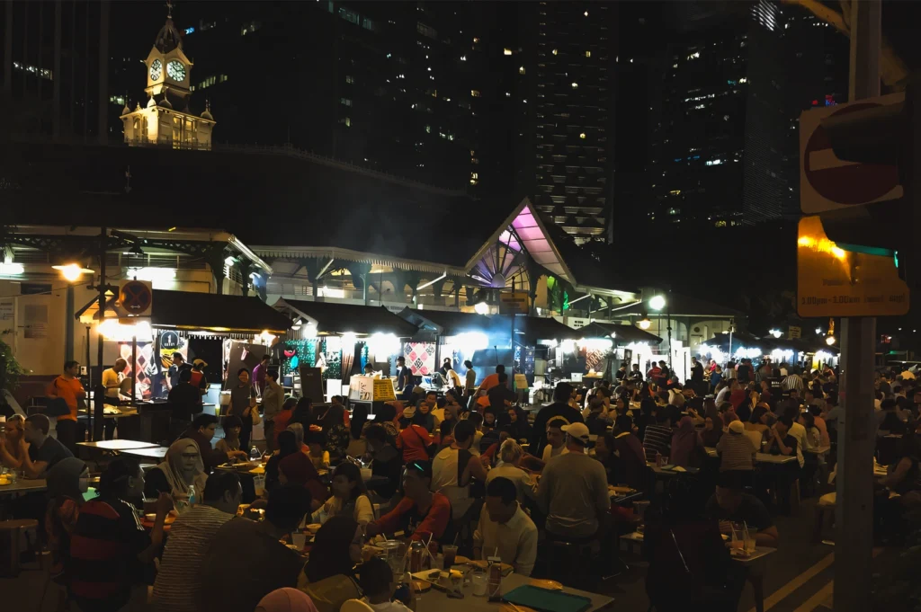 Wide‑angle night shot of a bustling street food market filled with outdoor dining tables, glowing food stalls, and city skyscrapers in the background, capturing vibrant urban nightlife and after‑hours dining culture.