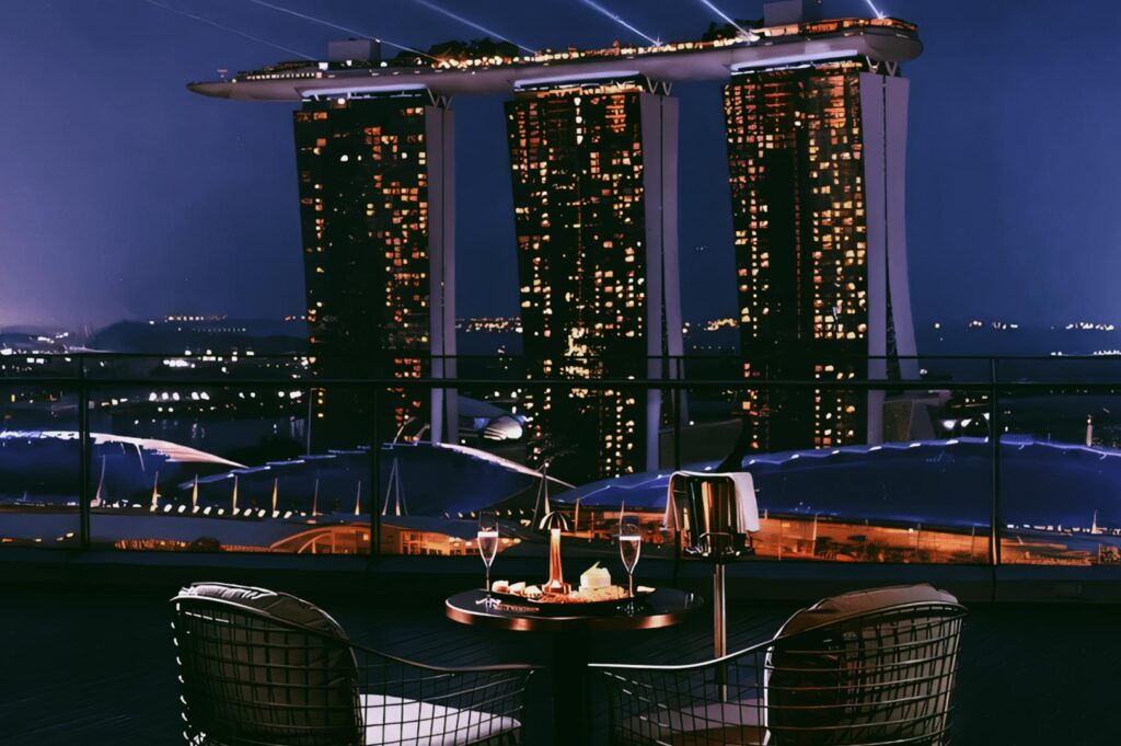 Romantic rooftop dining setup overlooking Marina Bay Sands at night, featuring candlelit wine glasses, a dessert platter, and panoramic views of Singapore’s illuminated skyline.
