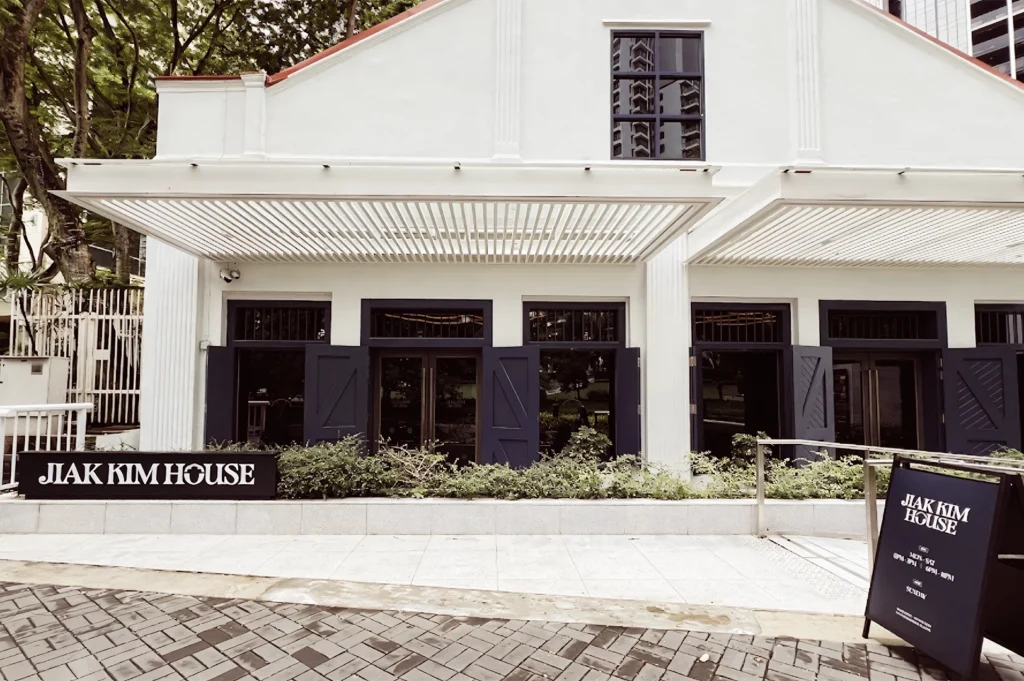 Front facade of Jiak Kim House with white colonial-style architecture, black-framed windows, and a walkway leading to the entrance surrounded by greenery.
