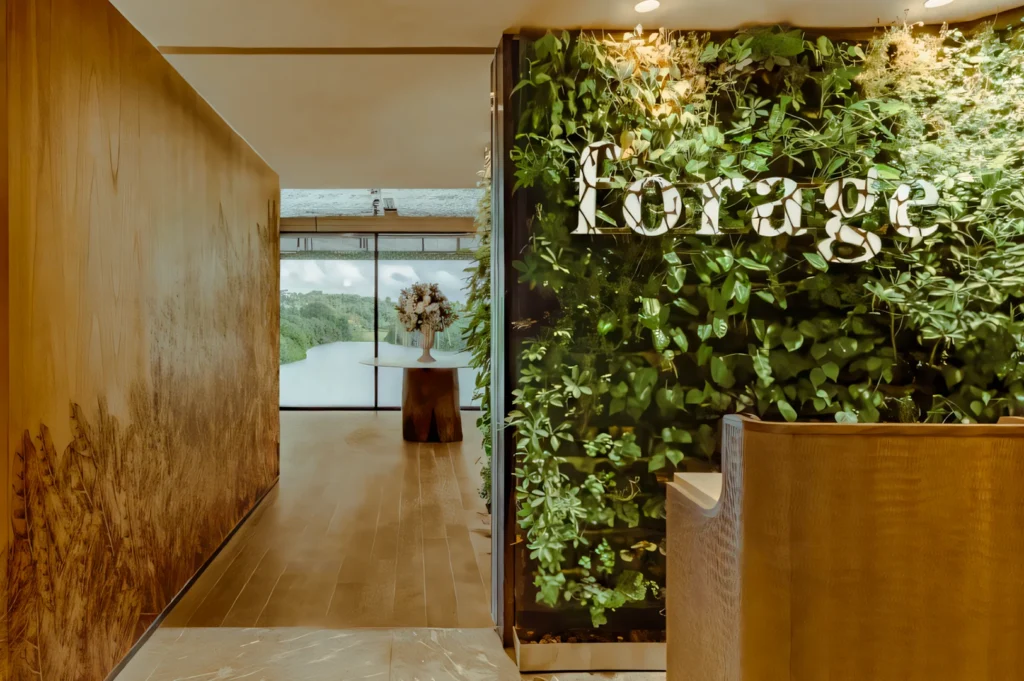 Wide‑angle eye‑level shot of a modern restaurant entrance featuring a vertical green wall with illuminated signage, wooden interior walls, and a minimalist reception area leading toward large windows with a scenic nature view.