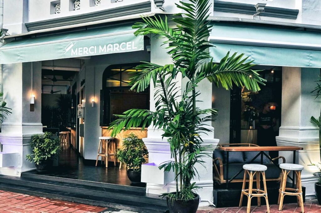 Exterior of Merci Marcel café with teal awnings, large potted plants, and outdoor seating in front of a white colonial-style building.