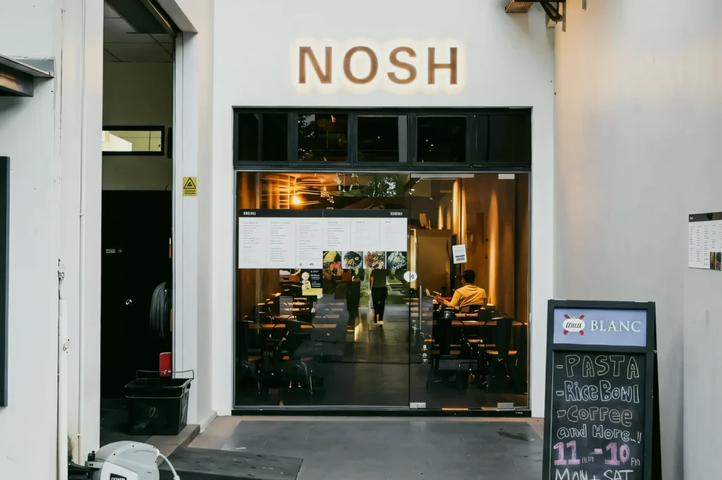 Eye-level straight-on shot of the NOSH restaurant storefront with glass entrance, illuminated signage, visible indoor seating, and a sidewalk menu board.