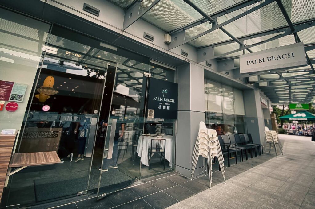Entrance of Palm Beach Seafood restaurant featuring a modern glass storefront, signage above the doorway, and outdoor seating stacked alongside the entrance.