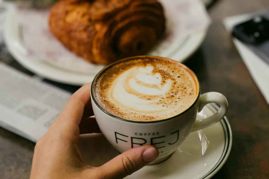 Close‑up eye‑level shot of a hand holding a cappuccino with latte art in a ceramic cup, paired with a flaky croissant on a café table, showcasing specialty coffee culture, café hopping, and relaxed brunch moments.