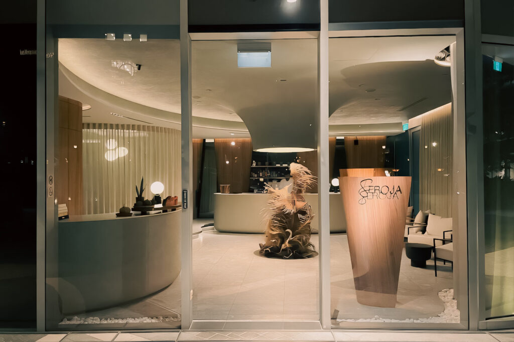 Elegant restaurant entrance with floor‑to‑ceiling glass windows, modern curved interior design elements, and the Seroja signage displayed on a wooden panel.