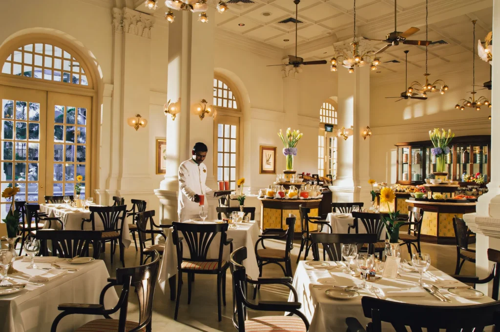 Wide‑angle interior shot of an elegant heritage restaurant with high ceilings, arched windows, white tablecloth dining tables, classic wooden chairs, and warm ambient lighting, showcasing Singapore’s colonial‑era dining atmosphere and refined culinary history.