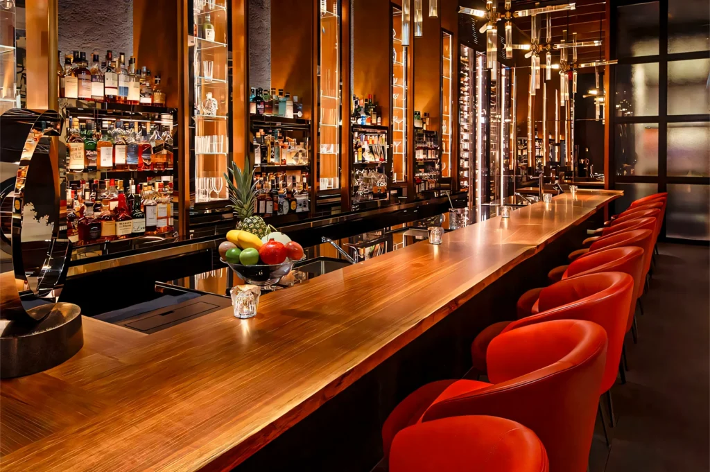 High‑end cocktail bar with a long polished wooden counter, red upholstered bar stools, and backlit shelves filled with premium spirits.