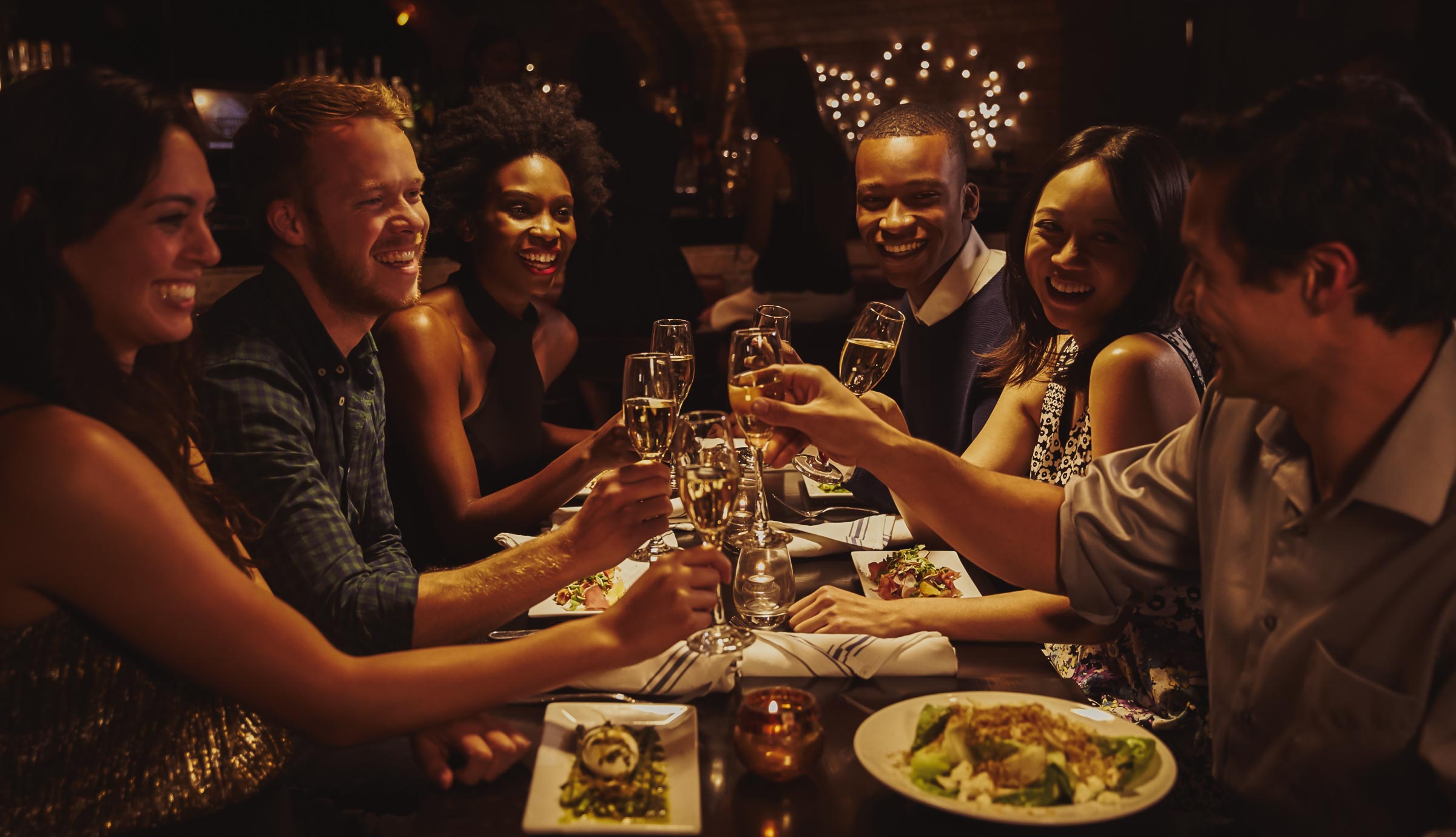 Diverse group of friends smiling and toasting with champagne glasses at a festive dinner party in a dimly lit restaurant.