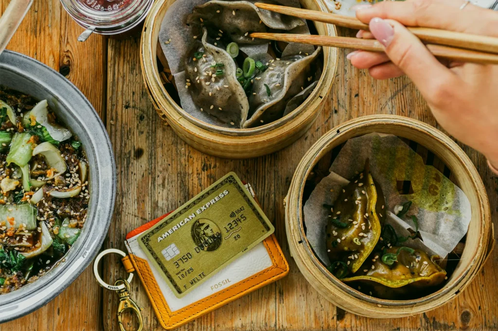 Top-down overhead shot of shared Asian dishes including dumplings on a wooden table, with chopsticks and payment cards, representing budget-friendly dining and meal deals.
