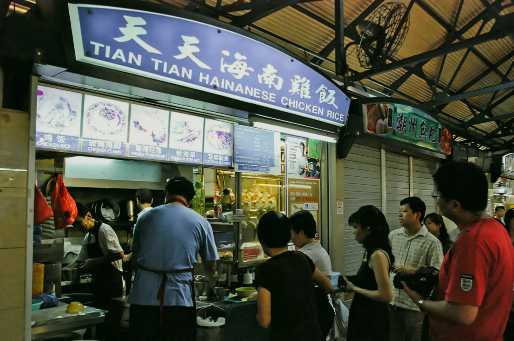 Eye-level wide-angle shot of Tian Tian Hainanese Chicken Rice hawker stall, showing customers queueing at a brightly lit food counter inside a covered market.