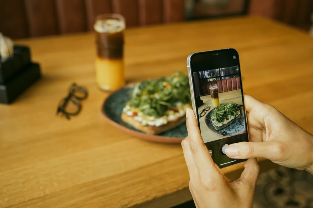 Over‑the‑shoulder close‑up shot of a person photographing a plated dish and coffee on a wooden restaurant table using a smartphone, highlighting dining as a shared food experience.
