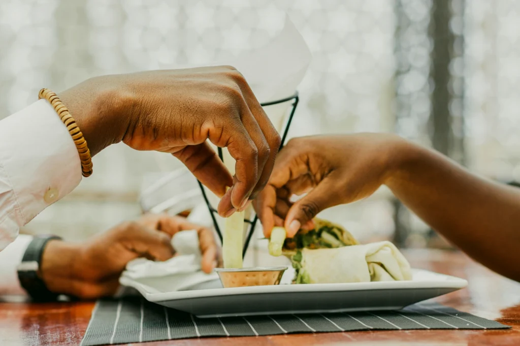 Eye-level close-up of two people sharing a meal at a restaurant, hands reaching toward a plate of food, capturing intimacy, connection, and dining as a love language.
