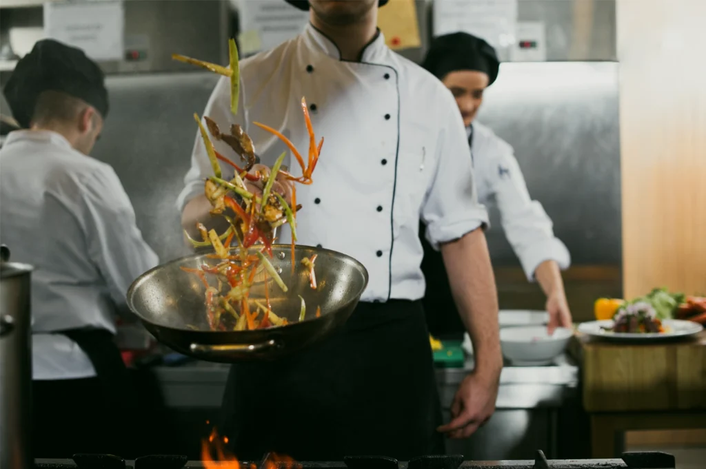 Medium shot at eye level of a professional chef tossing vegetables in a hot pan over an open flame inside a commercial restaurant kitchen.