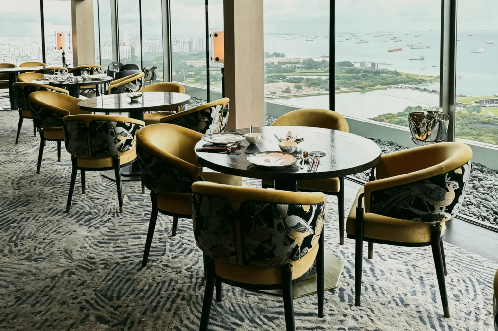 Wide‑angle interior shot of Jin Ting Wan Marina Bay Sands showing elegant round tables and Song Dynasty–inspired seating, with panoramic Marina Bay and Singapore waterfront views through floor‑to‑ceiling windows.