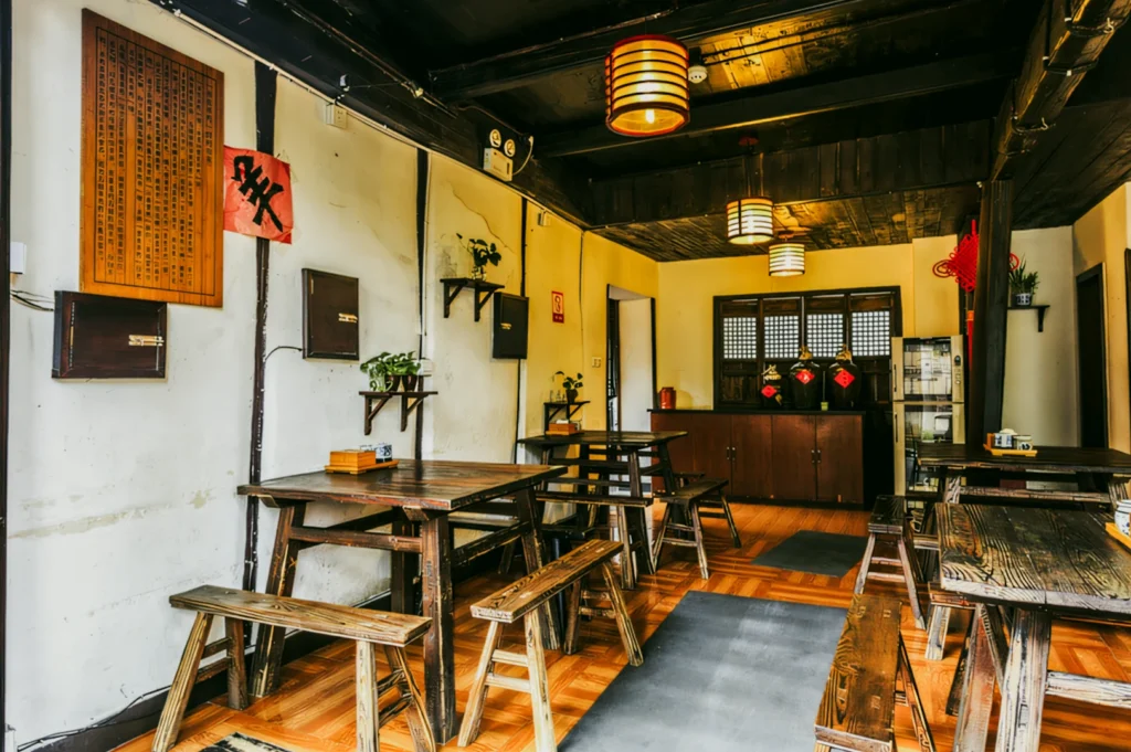 Wide-angle interior shot of a traditional-style restaurant dining space with rustic wooden tables and benches, warm hanging lantern lights, wooden flooring, and decorative wall accents, showcasing cultural ambiance and authentic restaurant design.