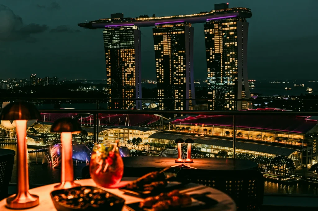 Eye‑level wide‑angle interior shot of a luxury restaurant dining table beside floor‑to‑ceiling windows overlooking Marina Bay Sands and Singapore harbor.