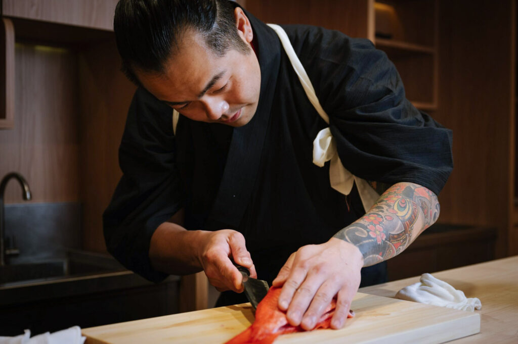Japanese chef filleting red fish on wooden board, tattoo sleeve visible, traditional black robe with white sash.