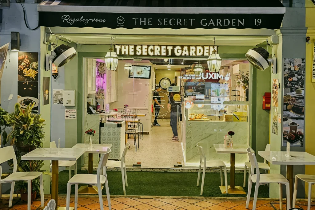 Straight-on street-level shot of “The Secret Garden” café storefront with green façade, outdoor white tables and chairs, warm interior lighting, and clear signage, presenting an inviting boutique café exterior at night.