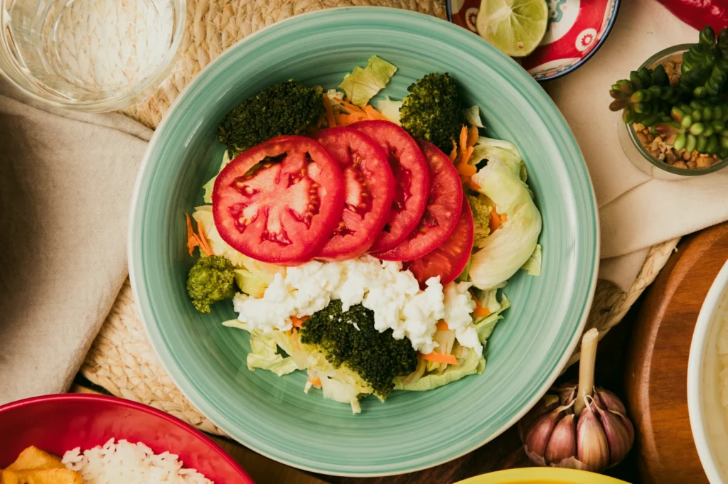 Overhead top‑down close‑up shot of a vegetarian salad bowl with sliced tomatoes, broccoli, lettuce, rice, and mixed vegetables arranged on a light blue plate on a rustic table setting.