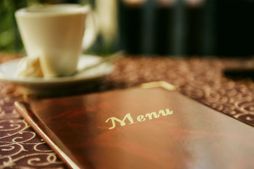 Shallow depth‑of‑field close‑up shot of a restaurant menu book on a patterned table, with the word “Menu” in focus and a cup of coffee softly blurred in the background.