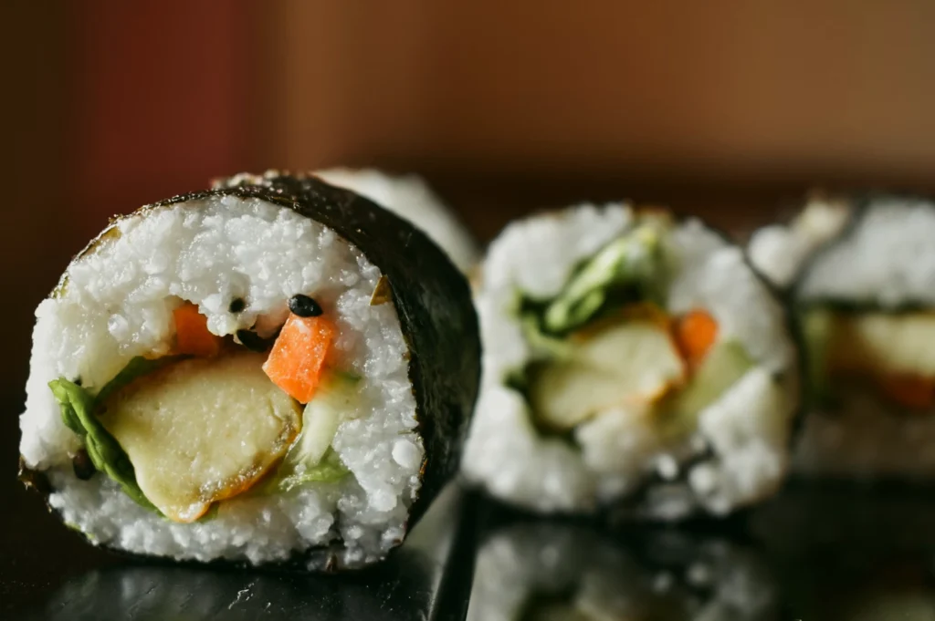 Macro close-up, eye-level shot of a sliced vegetable sushi roll wrapped in nori, showing white rice, avocado, carrot, and cucumber with shallow depth of field on a dark reflective surface.
