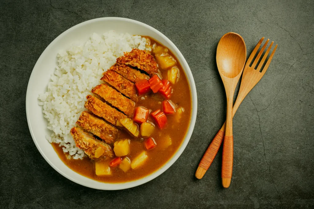 Top-down, flat-lay shot of a Japanese pork katsu curry rice dish served in a white bowl on a dark stone-textured surface, featuring neatly arranged white rice beside thick brown Japanese curry sauce with visible diced carrots and potatoes, topped with sliced golden-brown breaded pork cutlet. A wooden spoon and wooden fork rest parallel on the right side of the frame, with soft, even lighting highlighting the glossy curry texture and crisp panko coating, creating a clean, editorial food presentation.