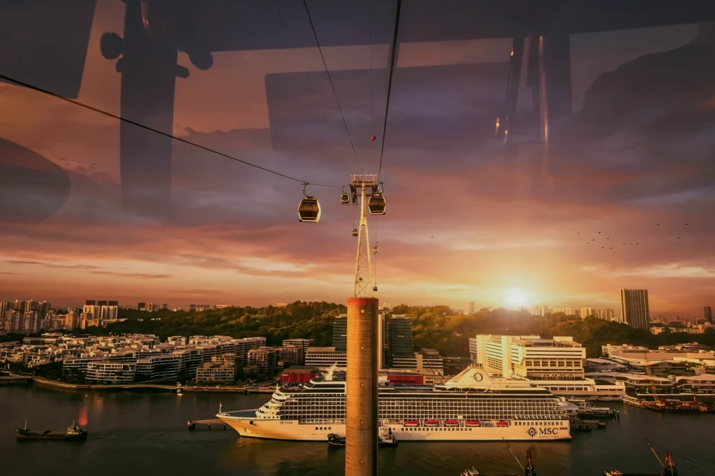 Wide-angle front-facing shot from inside Singapore cable car showing sunset skyline, Mount Faber Lodge aerial view, harbor cruise ship and cityscape – Cable Car Sky Dining experience Singapore.