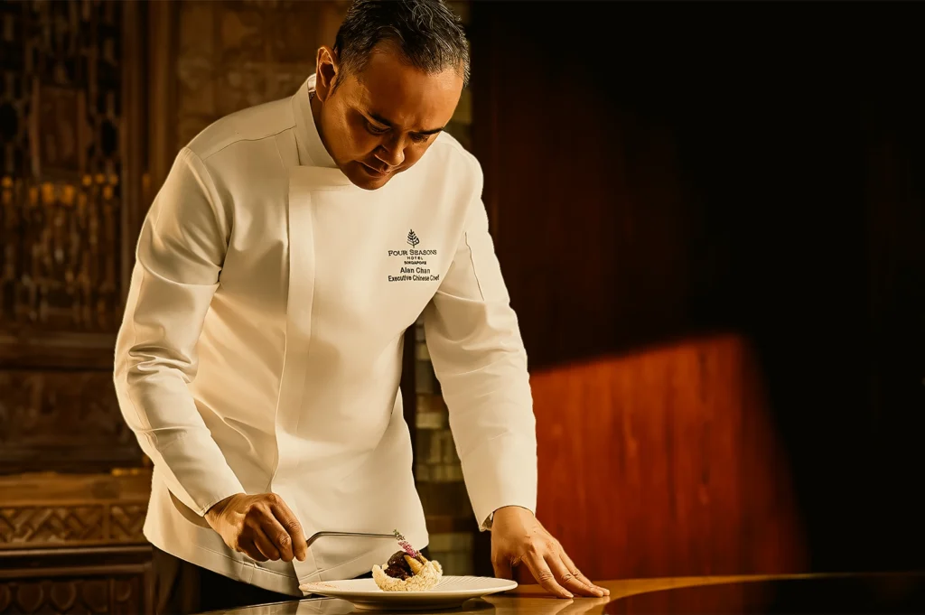 Medium close-up side-angle shot of professional chef plating gourmet dish at luxury Chinese restaurant in Singapore showcasing modern Cantonese cuisine
