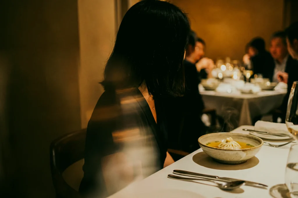 Eye-level shot of a refined fine dining restaurant scene with a diner seated beside a bowl of plated comfort food, warm ambient lighting and elegant table setting highlighting modern upscale cuisine.