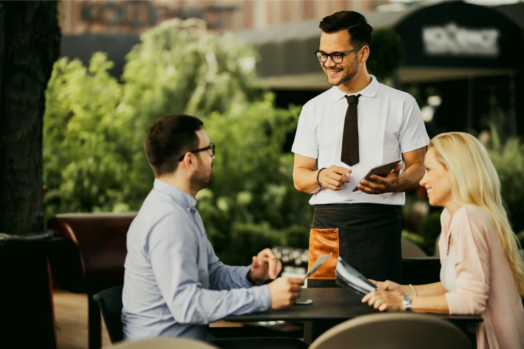 Eye-level medium shot of a server standing beside a café table while presenting a menu tablet to seated diners in an outdoor dining area surrounded by greenery, illustrating relaxed yet refined restaurant service.