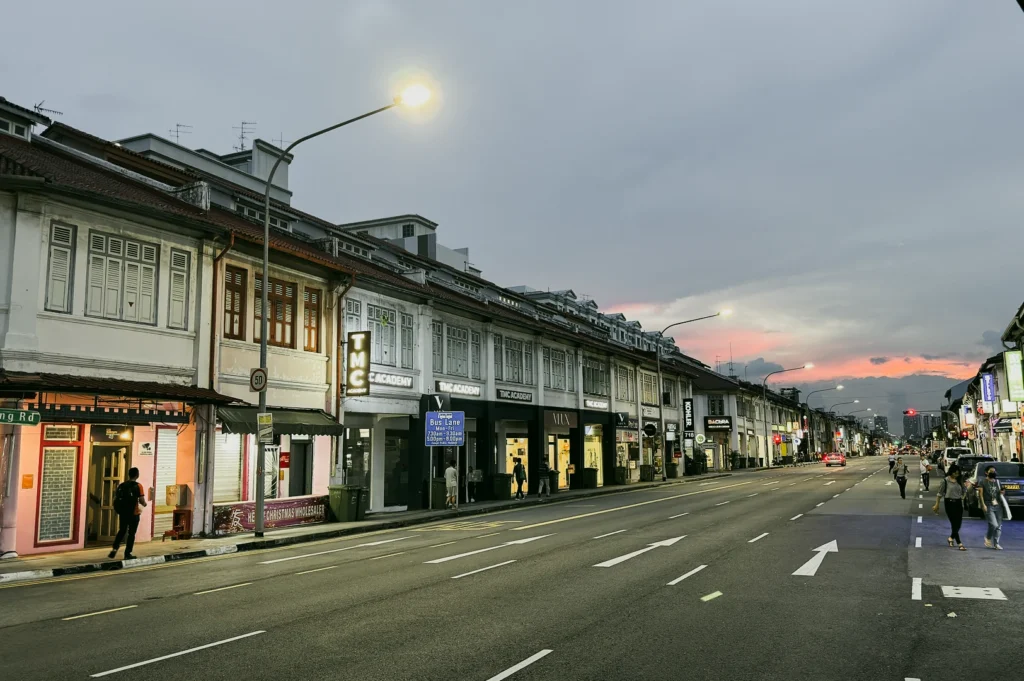 Heritage shophouses along Geylang Singapore at dusk with glowing streetlights and quiet road, shot at a low-angle leading-line perspective.