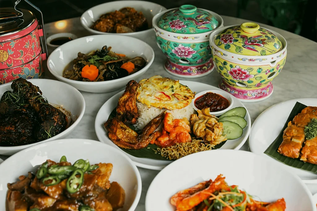 Eye-level slightly overhead shot of a Peranakan cuisine spread in Singapore featuring Nyonya dishes, rice with sambal, prawns, braised meats, and traditional ceramic serving ware on a marble table under warm lighting.