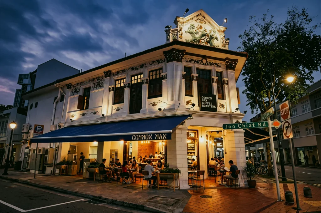 Low-angle wide shot of Common Man Coffee Roasters in Joo Chiat Singapore, a heritage shophouse café with outdoor dining, warm lighting, and neighborhood atmosphere.