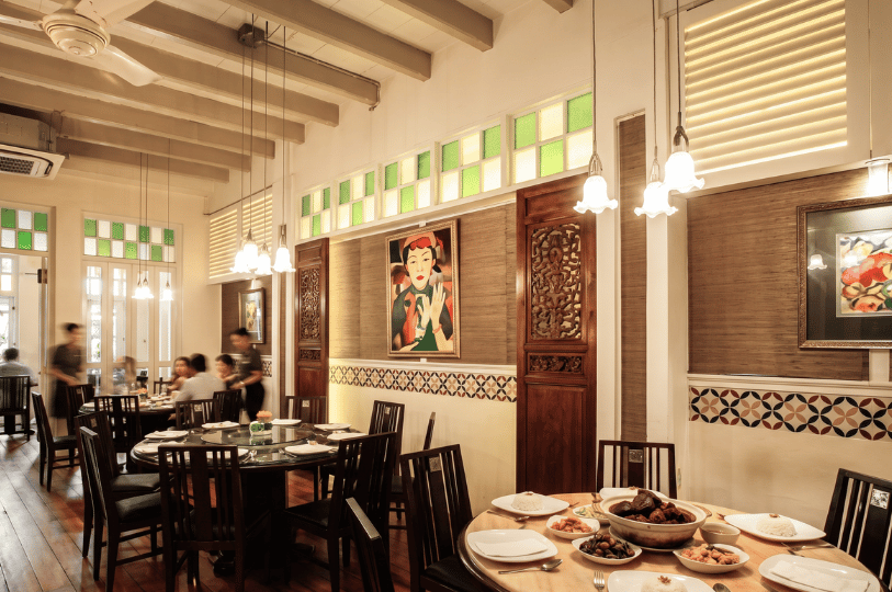 A warmly lit restaurant dining room with heritage-style decor, featuring exposed wooden ceiling beams, green and white stained-glass transoms, and a framed portrait painting on a textured wall. In the foreground, a round wooden table is set for a family-style meal with plates of white rice and several shared dishes. In the background, blurred diners enjoy a communal meal while a server attends to their table, evoking a nostalgic and welcoming atmosphere.