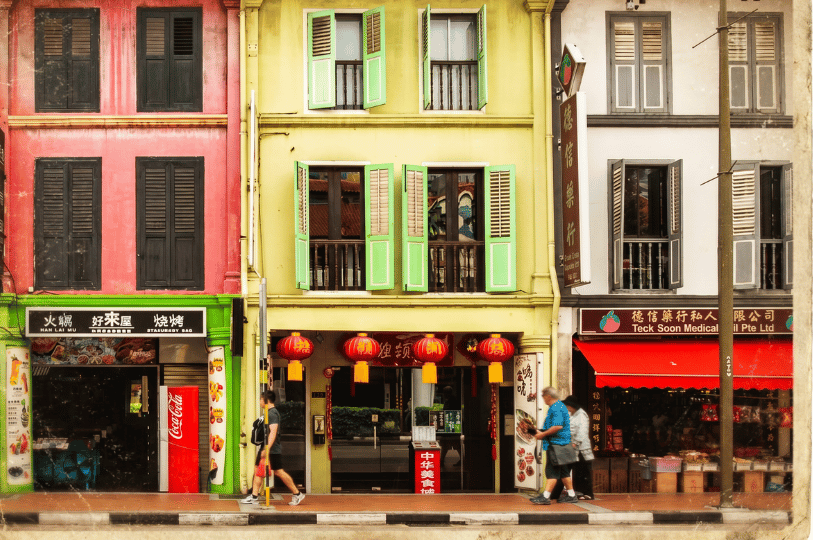 A row of brightly colored, historic shophouses featuring aging restaurants and traditional shops on the ground floor. The weathered but vibrant facades in deep pink, mustard yellow, and white—complete with open wooden shutters, hanging red lanterns, and classic storefront signage—capture the enduring, nostalgic charm of legacy dining neighborhoods.