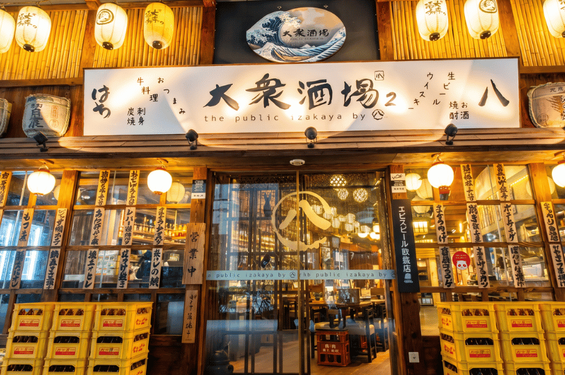 A warm, inviting wooden storefront of a Japanese izakaya, featuring glowing paper lanterns, traditional signage, stacked yellow crates, and glass doors that reveal a cozy, softly lit interior.
