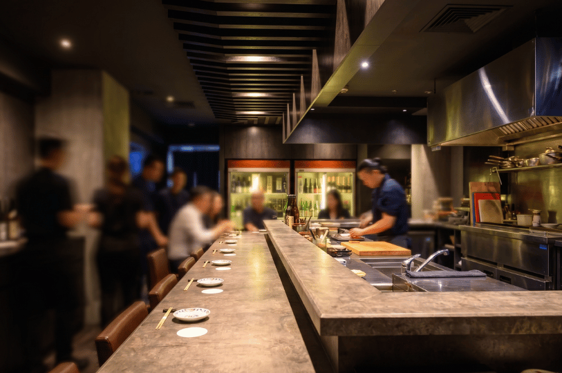 Interior of a Japanese restaurant featuring a long dining counter where patrons are seated while chefs prepare food in the open background kitchen.