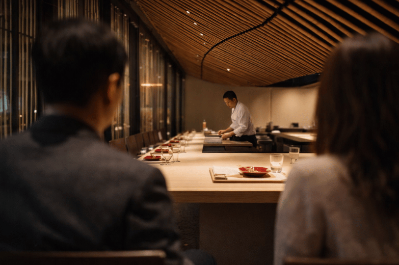 Diners sitting at a sleek, minimalist wooden omakase counter while a chef prepares a meal in the background under warm, elegant lighting.