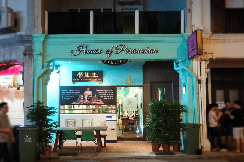 The exterior of House of Peranakan Petit at night, featuring a vibrant mint green shophouse facade, illuminated signage, and a small outdoor seating area with green chairs.